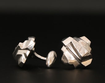 Puede incluir: Un par de gemelos de plata con un diseño geométrico. Los gemelos están hechos de un metal brillante y tienen una superficie texturizada.
