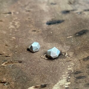 May include: A pair of silver geometric earrings, each shaped like a faceted gemstone. The earrings are small and delicate, and they are sitting on a wooden surface.