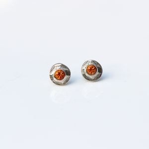 May include: A pair of silver stud earrings with a small orange gemstone in the center of each earring.