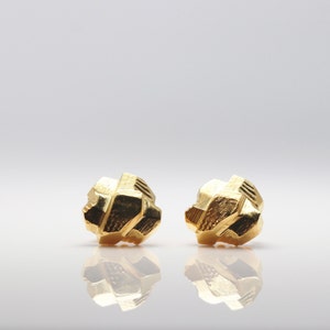 May include: A pair of gold-colored geometric stud earrings. The earrings are made of a textured metal and have a unique, abstract design.