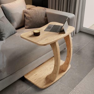 May include: A light wood C-shaped side table with a smooth rectangular top and a matching base, connected by a curved support. A silver laptop and a glass mug containing a dark liquid are placed on the surface of the table next to a sofa.