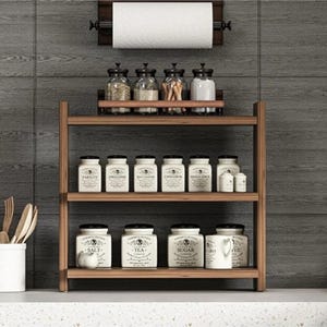 May include: A three-tiered wooden spice rack holding white ceramic canisters labeled with various spices and ingredients, with a wall-mounted paper towel holder above containing a roll of white paper towels, all set against a dark gray tiled wall.