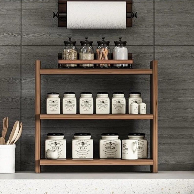 May include: A three-tiered wooden spice rack holding white ceramic canisters labeled with various spices and ingredients, with a wall-mounted paper towel holder above containing a roll of white paper towels, all set against a dark gray tiled wall.