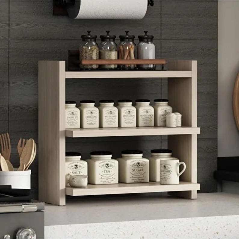 May include: A light wood two-tier spice rack featuring a top shelf with four glass jars and a bottom shelf holding several white ceramic canisters labeled with text including Salt, Tea, and Sugar, plus a white ceramic mug with a handle.