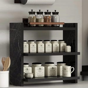 May include: A black marble-patterned spice rack with three tiers. The top shelf holds four clear glass jars with black lids. The middle shelf contains six white ceramic canisters with labels. The bottom shelf holds three white canisters and a white mug.