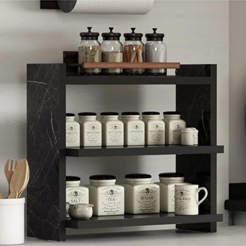 May include: A black marble-patterned spice rack with three tiers. The top shelf holds four clear glass jars with black lids. The middle shelf contains six white ceramic canisters with labels. The bottom shelf holds three white canisters and a white mug.