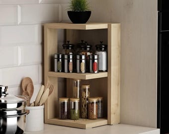 May include: A light-colored wooden corner shelf unit with three levels, holding various spice jars and a small potted plant. The shelf is filled with glass jars and metal spice containers, perfect for kitchen organization.
