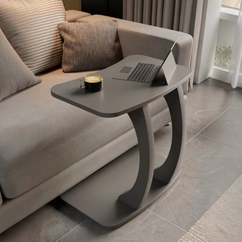 May include: A gray C-shaped side table with a smooth rectangular top surface. The table features a curved support base and holds a silver laptop computer and a small glass mug filled with a dark beverage, positioned next to a light gray sofa.