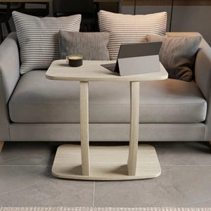 May include: A light wood C-shaped side table with a rectangular top and a matching rectangular base connected by two curved vertical supports. A dark gray tablet and a small black candle in a glass container are placed on the table surface.