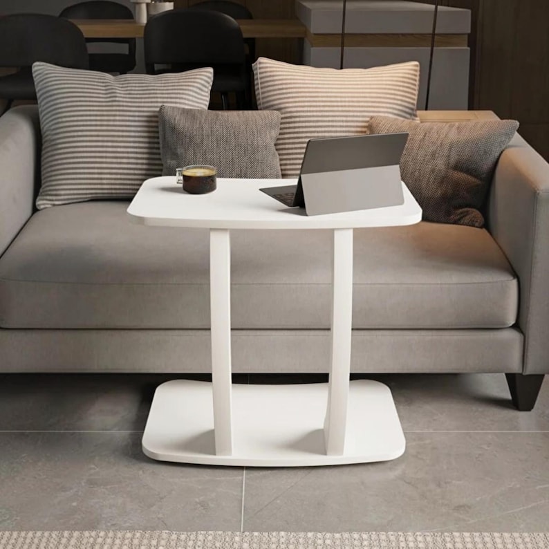 May include: A white C-shaped side table with a rectangular top and a flat base, featuring two vertical supports. A dark gray tablet and a small glass cup containing a dark liquid are placed on the surface of the table in front of a gray sofa.