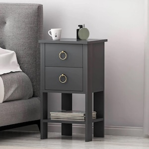 Pode incluir: Uma mesa de cabeceira cinza escuro com duas gavetas e puxadores de anel dourados. Uma prateleira abaixo contém revistas. Uma caneca branca e um frasco de perfume verde estão em cima. A mesa de cabeceira está ao lado de uma cabeceira cinza estofada.