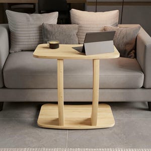 May include: A light-colored wooden side table with a rectangular top and base connected by two curved vertical supports. The table holds a dark gray tablet computer and a small glass cup containing a dark liquid, positioned in front of a sofa.
