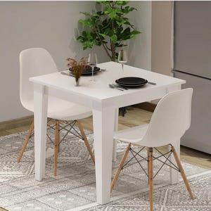 Könnte beinhalten: Ein weißer quadratischer Esstisch mit zwei weißen Stühlen. Die Stühle haben Holzbeine und eine Metallstützstruktur. Der Tisch ist mit schwarzen Tellern, Besteck und Weingläsern gedeckt. Eine dekorative Pflanze befindet sich im Hintergrund.