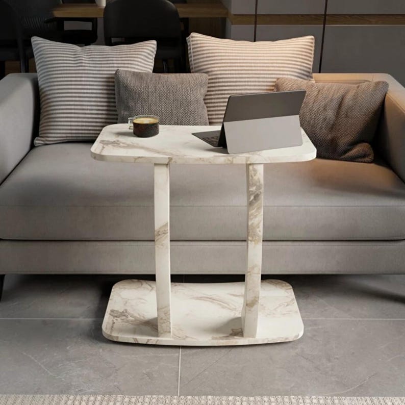 May include: A white marble-patterned side table with a rectangular top and base connected by two vertical supports. A dark gray tablet and a small glass mug containing a dark liquid are placed on the surface of the table.