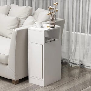 May include: A white bedside table with a small cabinet door and an open shelf. A white teacup and saucer sit on top, next to a vase with decorative stems. The table is next to a white sofa.