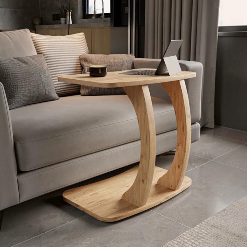 May include: A light-toned wooden C-shaped side table with a flat rectangular top and a curved support base. The table surface holds a dark-colored mug and a silver laptop computer. The structure is made of smooth wood with visible grain patterns.