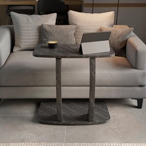 May include: A dark gray side table with a white marble-like vein pattern, featuring a rectangular top, two vertical supports, and a flat base. A small black cup and a silver-colored tablet computer are placed on the surface of the table.