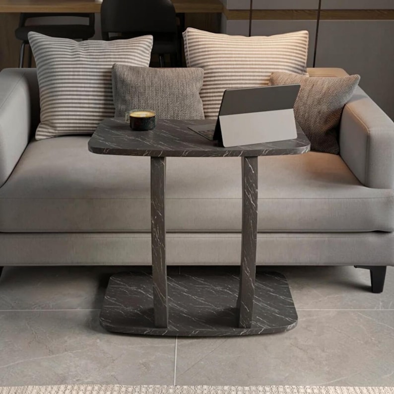 May include: A dark gray side table with a white marble-like vein pattern, featuring a rectangular top, two vertical supports, and a flat base. A small black cup and a silver-colored tablet computer are placed on the surface of the table.