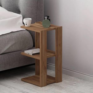 May include: A wooden side table with two shelves and a rectangular top. A white mug with a black design, a green bottle, and a book rest on the table. The table is next to a bed with a gray headboard.