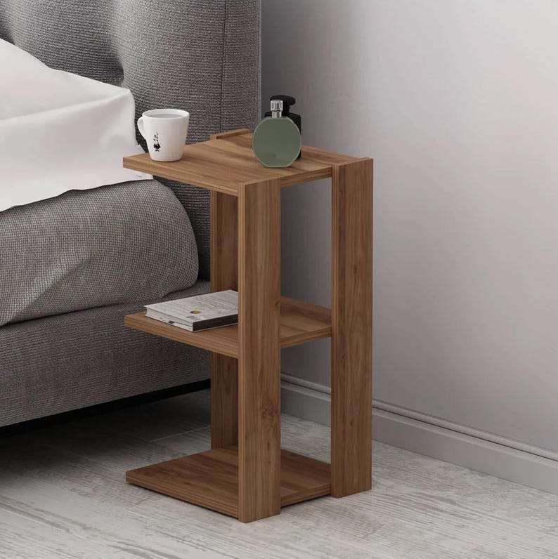May include: A wooden side table with two shelves and a rectangular top. A white mug with a black design, a green bottle, and a book rest on the table. The table is next to a bed with a gray headboard.