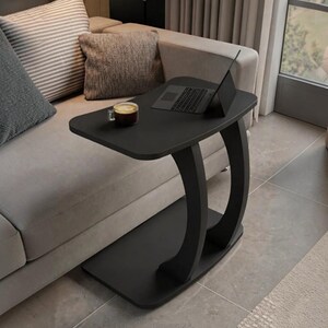 May include: A black C-shaped side table with a smooth rectangular top and a curved base support. The table features a flat surface holding a dark laptop and a small glass mug filled with a brown beverage, designed to slide over a sofa edge.