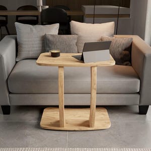 May include: A light-toned wooden C-shaped side table with a rectangular top and a matching rectangular base connected by two vertical supports. A dark gray tablet and a small dark mug are placed on the surface of the wooden table.