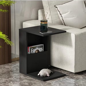 May include: A black side table with a magazine rack and a small shelf. A glass jar with a gold rim and a smartphone sit on the top shelf. A stuffed animal rests on the bottom shelf. A copy of Forbes magazine is in the rack.