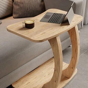 Può includere: Un tavolino laterale a forma di C in legno chiaro con un laptop e una tazza di caffè sopra. Il tavolo ha una base curva e un ripiano inferiore. Il tavolo è accanto a un divano grigio.