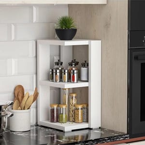 May include: A white two-tier corner shelf unit holding various glass spice jars with metal and wooden lids. A small potted green plant sits on the top shelf. Next to the shelf, a white ceramic container holds several wooden kitchen utensils.