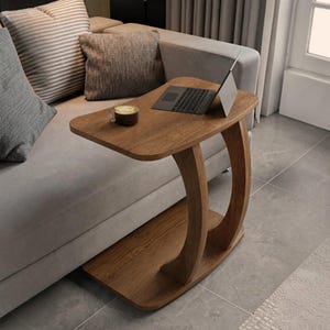 May include: A C-shaped side table with a wood grain finish, featuring a flat rectangular top and a curved support base. A silver laptop is open on the surface, positioned next to a small glass mug containing a dark brown beverage.