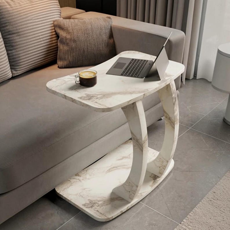 May include: A C-shaped side table with a white and gray marble-patterned finish. The table features a flat rectangular top and a matching base connected by a curved support structure. A laptop and a glass mug of dark liquid sit on the surface.