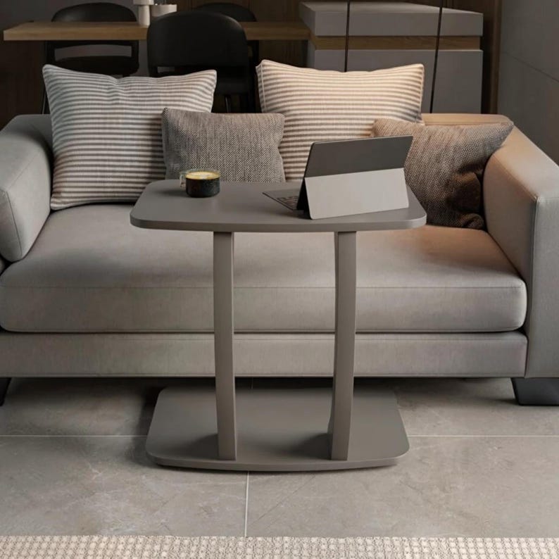 May include: A gray C-shaped side table with a rectangular top and a flat base. On the table surface sits a small dark mug and a gray tablet computer. The table is positioned in front of a light gray sofa with several striped and solid gray cushions.