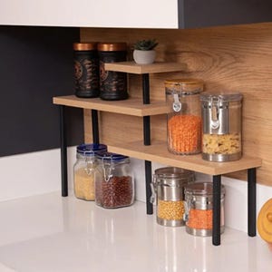 May include: A three-tiered wooden kitchen shelf with black metal legs holding various glass storage jars filled with grains, beans, and pasta, plus a small potted succulent on the top level. The jars feature metal latch lids.