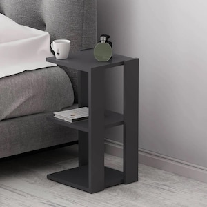 May include: A dark gray, modern side table with three shelves. A white mug, a green perfume bottle, and a book are on the shelves. The table is next to a gray bed.