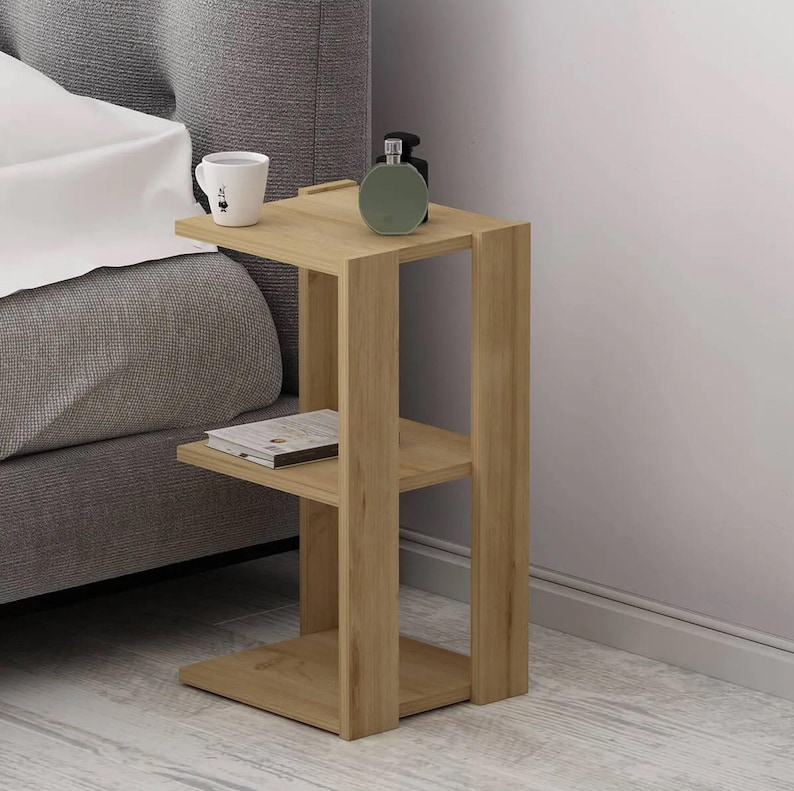 May include: A light brown wooden side table with three shelves. A white mug, a green bottle, and a book are on the shelves. The table is next to a gray bed.