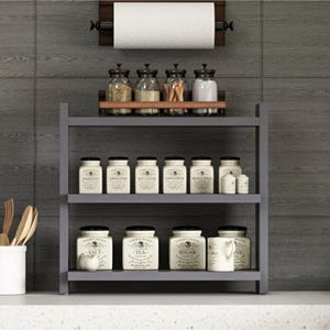 May include: A three-tiered gray kitchen shelf holding various glass spice jars with black lids on the top shelf, and white ceramic canisters labeled Salt, Tea, Sugar, and Coffee on the lower shelves, with a paper towel holder mounted above.