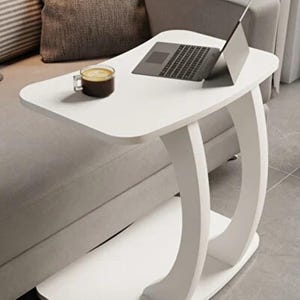 May include: A white, modern C-table with a curved base. A laptop and a cup of coffee sit on the tabletop. The table is positioned next to a gray sofa, suggesting a living room setting.
