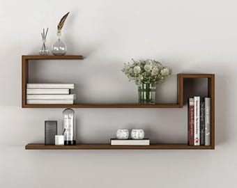 May include: A dark brown wooden wall shelf unit with two levels. The top shelf holds a glass vase with a feather, a stack of white books, and a glass vase with white flowers. The bottom shelf holds a glass cylinder, two white spheres, and books.