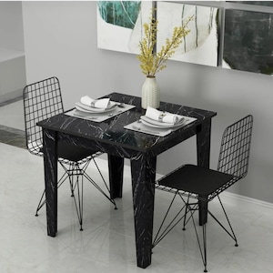 Op de afbeelding: Een vierkante eettafel met een zwart marmerpatroon blad en vier rechte poten, vergezeld door twee zwarte metalen draadstoelen. Een witte, getextureerde vaas met gele bloemen staat in het midden, geflankeerd door twee tafeldekkingen met witte borden.