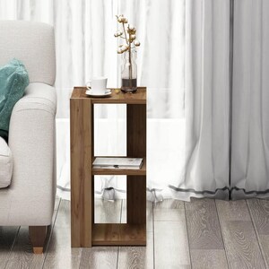 May include: A wooden side table with two shelves. A white teacup and saucer, a clear vase with cotton stems, and a book are on the table. The table is next to a white sofa and sheer curtains.