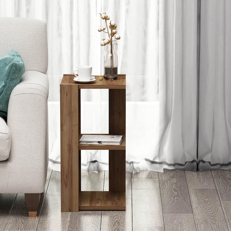 May include: A wooden side table with two shelves. A white teacup and saucer, a clear vase with cotton stems, and a book are on the table. The table is next to a white sofa and sheer curtains.