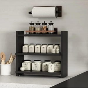 May include: A black wooden three-tier spice rack holding white ceramic jars with black text labels. Above the rack, a wall-mounted paper towel holder with a roll of white paper towels. A white utensil holder containing wooden spoons sits nearby.