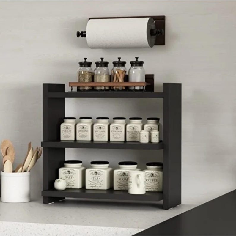 May include: A black wooden three-tier spice rack holding white ceramic jars with black text labels. Above the rack, a wall-mounted paper towel holder with a roll of white paper towels. A white utensil holder containing wooden spoons sits nearby.