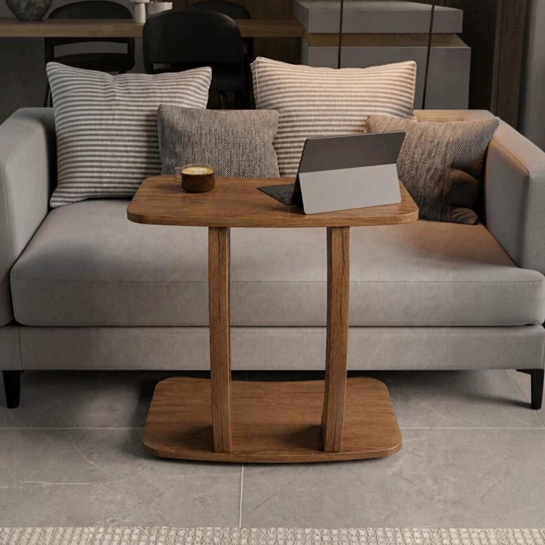 May include: A wooden C-shaped side table with a rectangular top and base, featuring two vertical supports. A small brown mug and a grey tablet are placed on the surface. The table is positioned in front of a light grey sofa with striped and solid cushions.