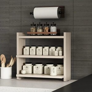 May include: A light wood three-tiered countertop spice rack holding white ceramic canisters labeled with spice names, with a matching paper towel holder mounted above, and a white utensil crock containing wooden spoons on the side.
