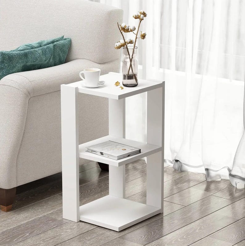 May include: A white, modern side table with a three-tiered design. The table has a square top and two shelves, perfect for displaying decor. A coffee cup, a vase with cotton stems, and a magazine are on the table. The table is next to a white sofa.