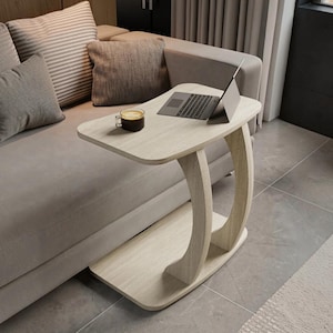 May include: A light wood C-shaped side table with a flat rectangular top and a curved support base. A dark gray laptop is open on the surface, positioned next to a clear glass mug containing a dark brown beverage with a light foam top.