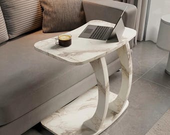May include: A C-shaped side table with a white and gray marble-patterned finish. The table features a flat rectangular top and a matching base connected by a curved support structure. A laptop and a glass mug of dark liquid sit on the surface.