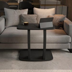 May include: A black C-shaped side table with a rectangular top and a flat base. On the table surface sits a dark gray tablet in a stand and a small glass cup filled with a dark brown liquid. The table is positioned in front of a gray sofa.