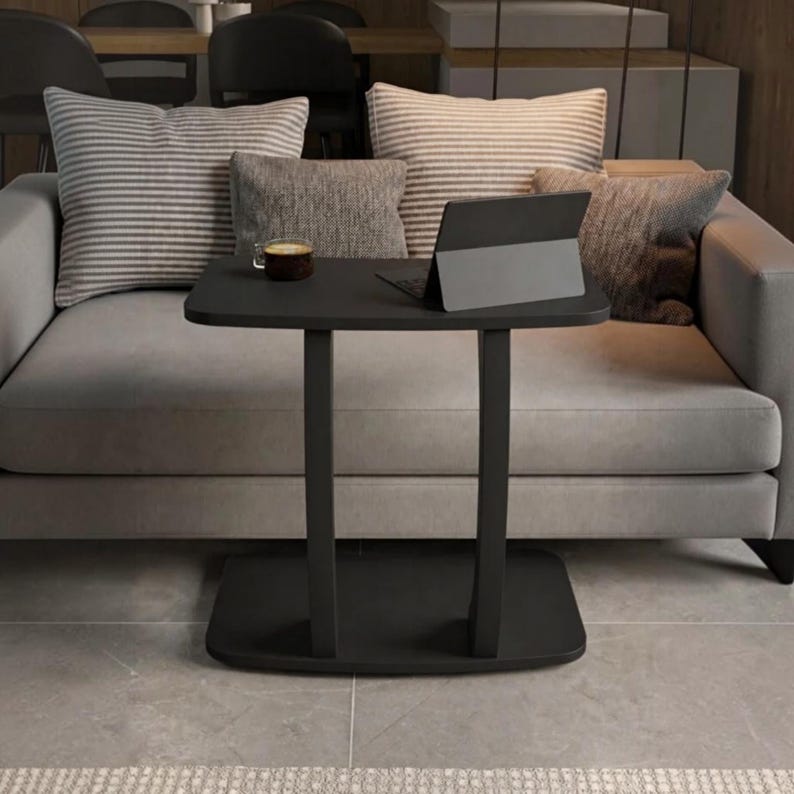 May include: A black C-shaped side table with a rectangular top and a flat base. On the table surface sits a dark gray tablet in a stand and a small glass cup filled with a dark brown liquid. The table is positioned in front of a gray sofa.
